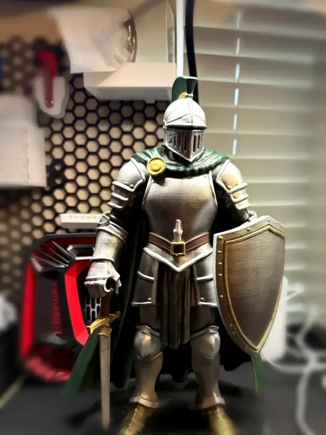 Painted knight mascot statue from the front, with silver armor, a green cape and plume, gold boots, a sword, and a shield on a stepped base.